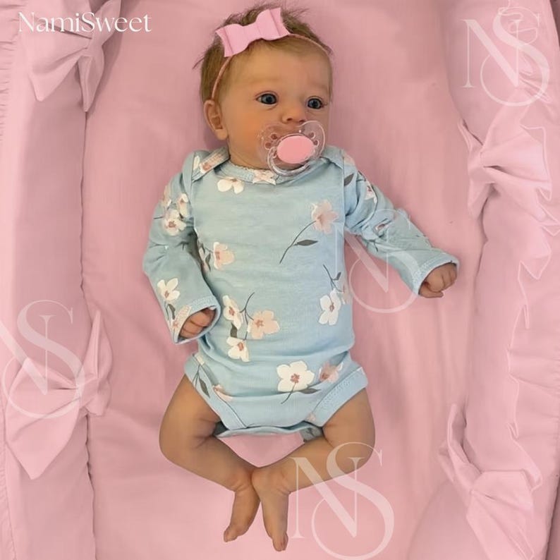 May include: A baby wearing a light blue floral onesie and a pink bow headband. The baby is lying on a pink surface and has a pink pacifier. The image has the text "NamiSweet" in the upper left corner.