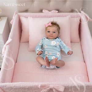 May include: A baby in a light blue floral onesie and pink headband rests in a pink crib. The crib has a quilted bumper, pillows, and a bow detail. The brand name "NamiSweet" is visible in the top left corner.