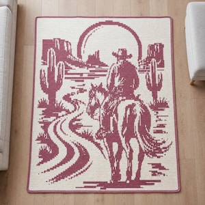 May include: A rectangular crochet rug featuring a two-tone design in cream and deep red. The pattern depicts a desert landscape with a large sun, cacti, rock formations, and a person on horseback riding along a winding path toward the horizon.