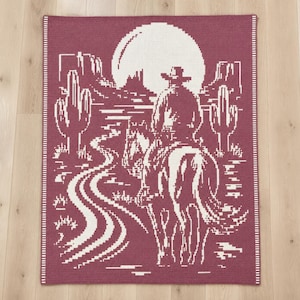 May include: A rectangular woven blanket featuring a deep maroon background with a white pixelated design. The pattern depicts a lone rider on horseback traveling along a winding path toward a large full moon, flanked by desert cacti and mesas.