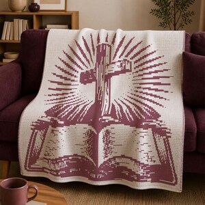 May include: A cream-colored throw blanket featuring a dark maroon pixelated graphic of an open book with a cross rising from the center, surrounded by radiating sunburst lines. The blanket has a textured waffle-knit pattern throughout the fabric.