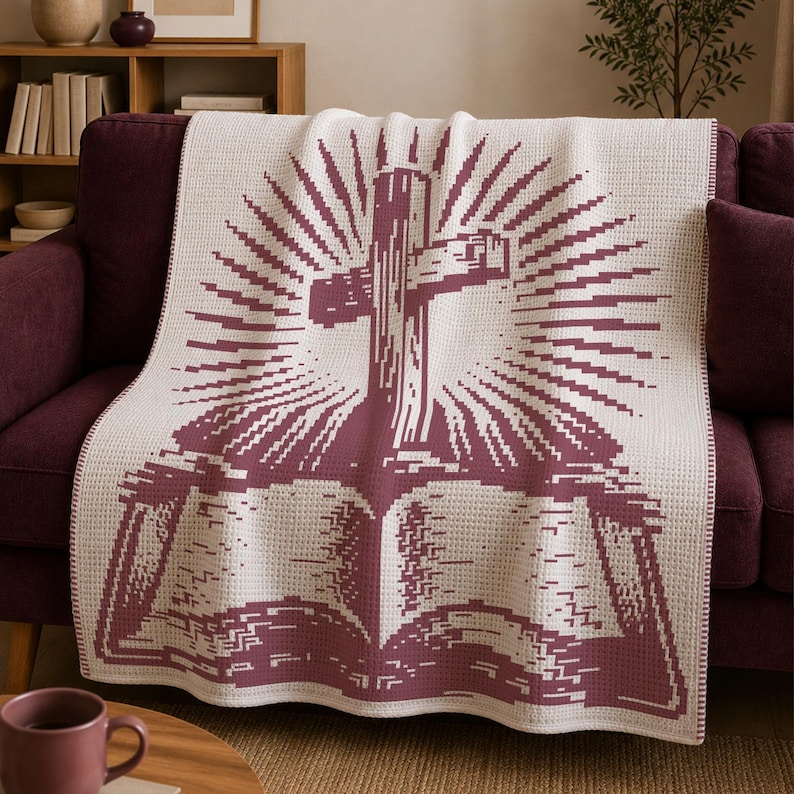 May include: A cream-colored throw blanket featuring a dark maroon pixelated graphic of an open book with a cross rising from the center, surrounded by radiating sunburst lines. The blanket has a textured waffle-knit pattern throughout the fabric.