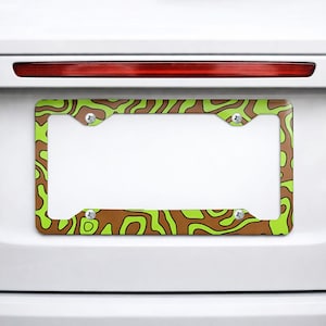 May include: A rectangular license plate frame featuring a vibrant pattern of irregular, flowing lime green and brown shapes. The frame has four circular mounting holes, each secured with a silver-toned metal screw, and is mounted on a white vehicle.