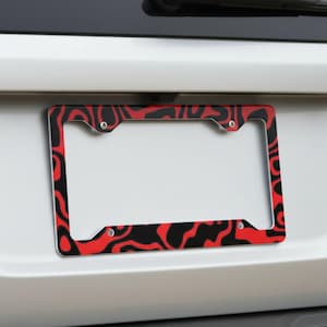 May include: A rectangular license plate frame featuring a bold, abstract pattern of flowing, organic shapes in vibrant red and deep black. The frame is secured with four visible silver-toned screws at the corners, creating a high-contrast design.