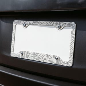 Op de afbeelding: Een rechthoekig kentekenplaathouder met een zwart-wit topografisch kaartpatroon van lijnen. Het frame is voorzien van vier ronde montagegaten, elk vastgezet met een zilverkleurige metalen schroef, gemonteerd op een donkere ondergrond.