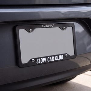 May include: A black license plate frame mounted on a gray vehicle bumper. The frame features the white text SLOW CAR CLUB at the bottom and Japanese characters at the top. Four silver screws secure the frame to the vehicle's rear bumper area.