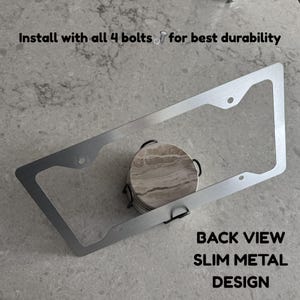 Op de afbeelding: Een slank, rechthoekig kentekenplaathouder van zilverkleurig metaal met vier montagegaten. De tekst op de afbeelding luidt: Install with all 4 bolts for best durability en BACK VIEW SLIM METAL DESIGN. Het frame ligt over een ronde stenen onderzetter.