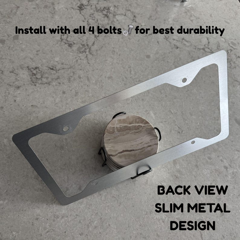 Op de afbeelding: Een slank, rechthoekig kentekenplaathouder van zilverkleurig metaal met vier montagegaten. De tekst op de afbeelding luidt: Install with all 4 bolts for best durability en BACK VIEW SLIM METAL DESIGN. Het frame ligt over een ronde stenen onderzetter.
