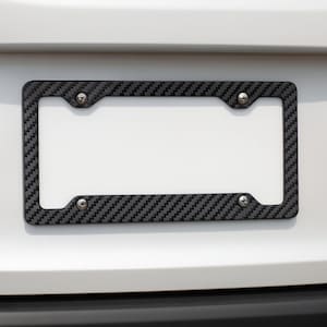 May include: A black and gray carbon fiber license plate frame with four silver screws. The frame has a textured, woven pattern and is mounted on a white surface.