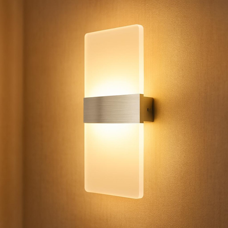 K&ouml;nnte beinhalten: Moderne Wandleuchte mit rechteckigem Schirm aus Milchglas. Ein geb&uuml;rstetes Metallband akzentuiert die Mitte, und die Leuchte ist auf einer quadratischen Metallbasis montiert. Das Licht strahlt ein warmes, einladendes Leuchten aus, ideal f&uuml;r die Innenbeleuchtung.