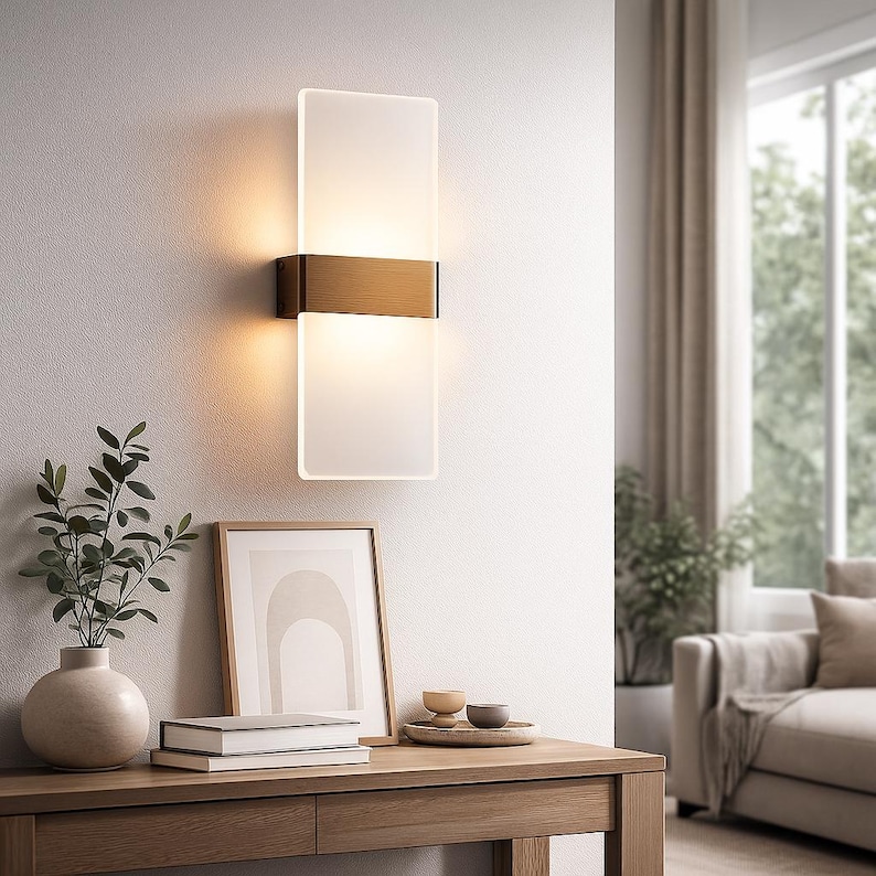K&ouml;nnte beinhalten: Moderne Wandleuchte mit rechteckigem, mattiertem Glasschirm, unterteilt durch ein horizontales Band in Bronzefarbe. Die Leuchte ist an einer wei&szlig;en Wand montiert und wirft ein warmes Licht. Das Design ist minimalistisch und elegant, geeignet f&uuml;r Innenbeleuchtung.