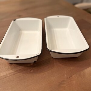 May include: Two rectangular loaf pans with a white enamel finish and a thin black rim. Each pan features a small hole on the handle tab at one end, designed for hanging. The interior and exterior surfaces are smooth, white, and glossy.