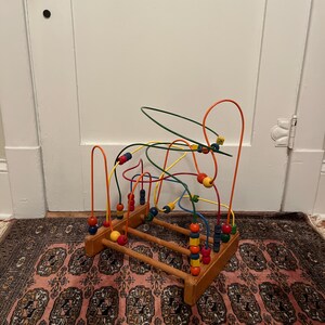 May include: A wooden bead maze toy with colorful beads and wire tracks. The base is made of wood, and the wires are bent into loops and curves. The beads are various colors, including red, yellow, blue, and green. The toy is designed for children.