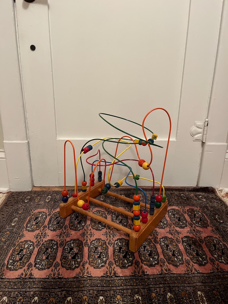 May include: A wooden bead maze toy with colorful beads and wire tracks. The base is made of wood, and the wires are bent into loops and curves. The beads are various colors, including red, yellow, blue, and green. The toy is designed for children.