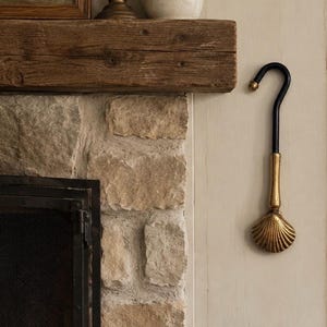 May include: A wall-mounted hook featuring a black curved metal top, a gold-toned cylindrical neck, and a decorative gold-toned seashell-shaped base. The hook is mounted on a light-colored wall next to a stone fireplace and a wooden mantelpiece.