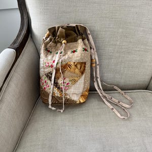 May include: A drawstring bag featuring a patchwork design with floral patterns, tan fabric, and brown accents. The bag has a solid brown top section, a long patterned strap, and wooden beads on the drawstring cords for closure.