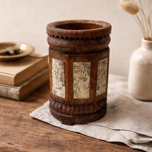 May include: A cylindrical wooden container with carved details and inset panels. The panels feature illustrations of figures in a cream color. The container is brown with a decorative rim and base. It sits on a wooden surface.