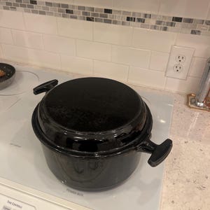 May include: A black cast iron Dutch oven with a lid, sitting on a white electric stovetop. The pot has two side handles and a black knob on the lid. The stovetop controls are visible.