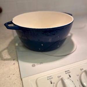 May include: A deep, round, dark blue enameled cast iron Dutch oven with two side handles and a smooth, cream-colored interior. The pot is positioned on a white GE Profile electric stovetop with visible control knobs and burner settings.