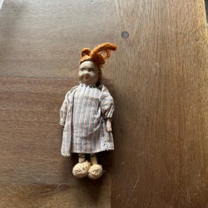 May include: A small antique doll with red yarn hair and a striped dress in shades of tan, blue, and white. The doll has crocheted shoes and a painted face. The doll is standing on a wooden surface.