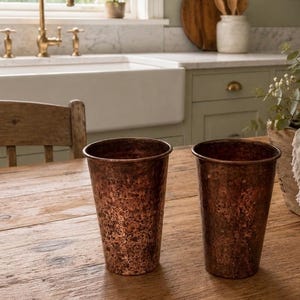 May include: Two copper-colored, hammered metal tumblers sit on a wooden table. The tumblers have a textured surface and a slightly tapered shape. The background includes a kitchen sink, faucet, and a wooden chair.