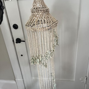 May include: A decorative hanging shell chandelier. The chandelier is made of various sizes of white and beige shells, strung together to form a conical shape. Strands of shells cascade downwards, with clusters of green shells interspersed throughout.