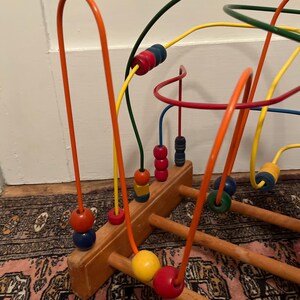 May include: A wooden bead maze toy with colorful, curved wires and wooden beads. The beads are various colors, including red, blue, yellow, and green. The base is made of light brown wood. The toy is designed for children.