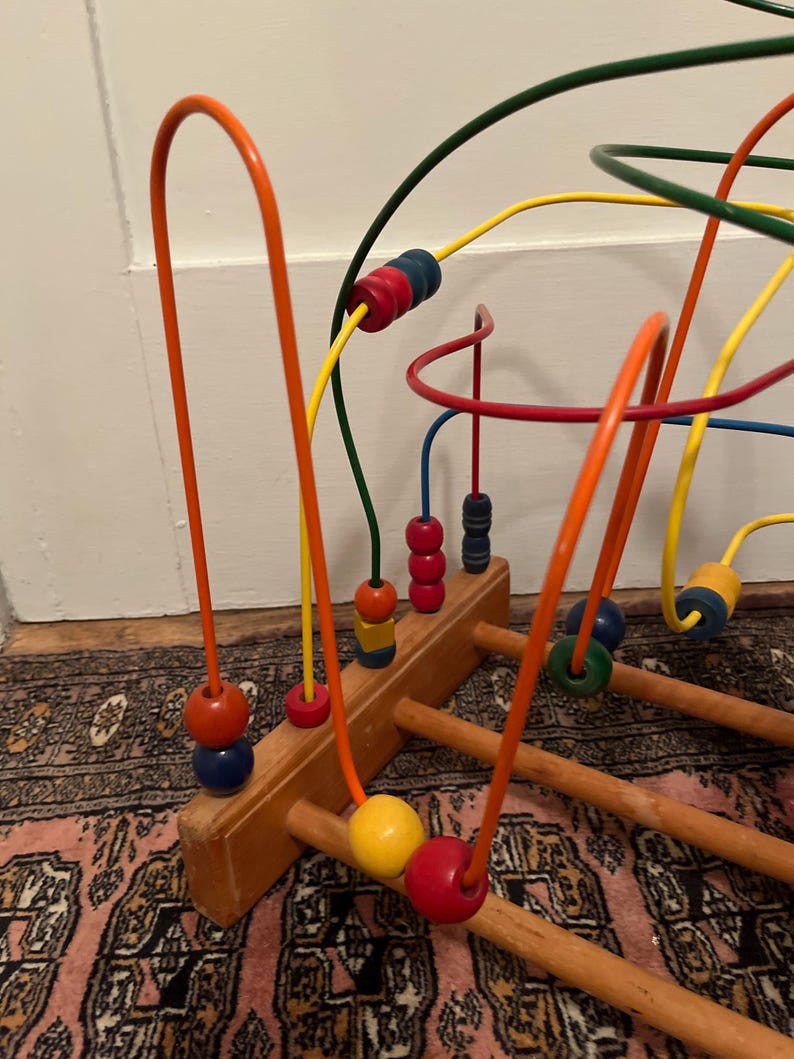 May include: A wooden bead maze toy with colorful, curved wires and wooden beads. The beads are various colors, including red, blue, yellow, and green. The base is made of light brown wood. The toy is designed for children.