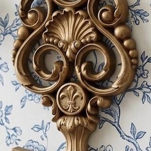 May include: A large, ornate gold-colored wall decoration shaped like a skeleton key, featuring intricate scrollwork, a central fleur-de-lis emblem, and a shell-like motif at the top, suspended from a matching gold-toned metal wall hook.