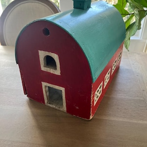 May include: A red wooden barn with a teal roof and a small chimney. The barn has white window and door frames. The barn is sitting on a wooden surface.