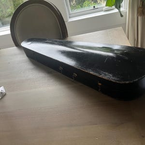 May include: A black, rectangular violin case with a slightly distressed finish. The case is closed and rests on a light-colored wooden table. The case has metal clasps and hinges. A clear glass candlestick is visible in the foreground.
