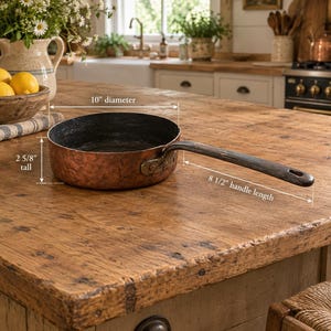 May include: A round copper pan with a dark interior and a long, dark metal handle. The pan features a 10-inch diameter, a height of 2 5/8 inches, and an 8 1/2-inch handle length. The exterior surface has a hammered texture and a metallic finish.