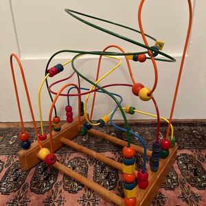 May include: A wooden bead maze toy with colorful, curved wires and wooden beads. The beads are various colors, including red, yellow, blue, and green. The base is made of natural wood.