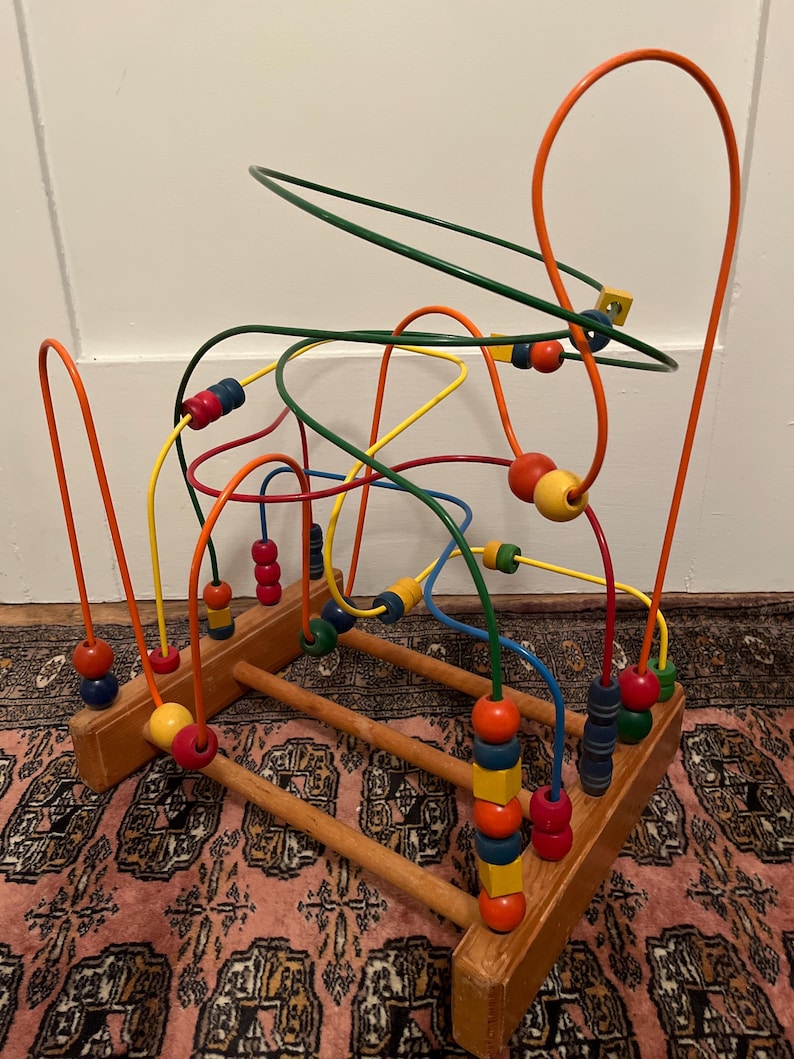 May include: A wooden bead maze toy with colorful, curved wires and wooden beads. The beads are various colors, including red, yellow, blue, and green. The base is made of natural wood.