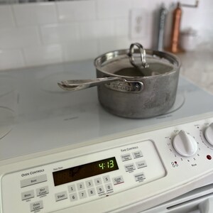 May include: A stainless steel saucepan with a matching lid sits on a white electric stove. The stove features a digital display showing 4:13, a numeric keypad, and control knobs. The saucepan has a long handle and the All-Clad brand name engraved.