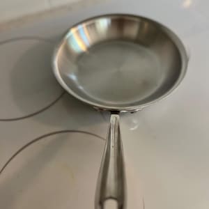 May include: A round stainless steel frying pan with a polished metallic finish and a long, slender handle featuring a circular hole at the end for hanging. The pan has a smooth, reflective interior surface and a sturdy, riveted handle construction.