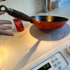 May include: An orange cast iron skillet with a dark interior and a wooden handle. A red Coca-Cola can sits next to the pan on a white electric stovetop. The stovetop has the words "HOT SURFACE" printed on it.