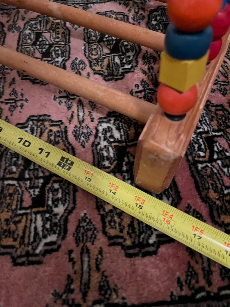 May include: Wooden abacus with colorful beads, including red, blue, and yellow. A yellow measuring tape shows a length of 16 inches. The abacus is on a pink patterned rug.