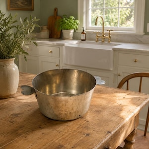 May include: A round, metallic basin with a weathered, silver-toned finish and two distinct heart-shaped handles on opposite sides. The bowl features a wide, open top and a flat base, displaying a textured, aged patina across its exterior surface.