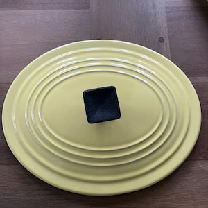 May include: A pale yellow, oval-shaped pot lid with a black square handle. The lid has concentric ridges and is set against a dark wood surface. This kitchenware item is designed for cooking.