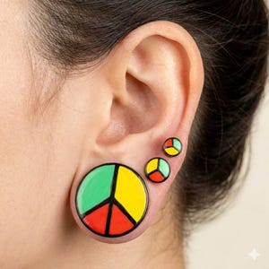 May include: A set of three ear gauges with a peace sign design. The largest gauge is green, yellow, and red, with a black outline. The smaller gauges are also in the same color scheme. The gauges are worn in the ear.
