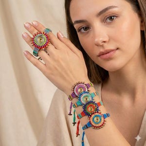 Puede incluir: Una colección de coloridas joyas de macramé, que incluyen un anillo y varias pulseras. El anillo y las pulseras presentan un diseño circular con vibrantes tonos de rojo, azul, verde y amarillo, acentuados con cuentas doradas.