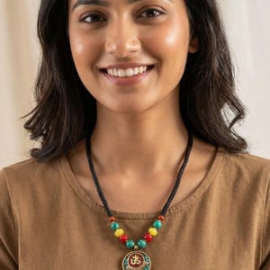 May include: A black cord necklace with colorful beads and a circular pendant. The pendant has a gold-colored border and a central design with red and gold accents. The necklace features red, yellow, and turquoise beads.