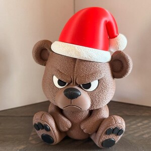 May include: A brown bear figurine with an angry expression, wearing a red Santa hat with a white trim. The bear is sitting with its paws out, and has black paw pads. The figurine is a Christmas decoration.