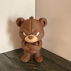 May include: A brown bear figurine with an angry expression, arms crossed. The bear has a textured surface and a tan snout. The figurine is standing on a dark wooden surface.