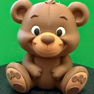 May include: A brown teddy bear figurine with a smiling face, large ears, and paw details. The bear has a light brown snout and paw pads, with black eyes and white highlights. The bear is sitting on a dark surface against a green background.