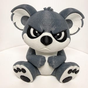 May include: A gray koala figurine with an angry expression. The koala has white accents on its ears, chest, and feet. Its eyes are black with white highlights, and it has a black nose. The figurine is sitting down.