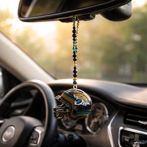 May include: A decorative car charm featuring a miniature football helmet with the Jacksonville Jaguars logo. The helmet is black with teal, gold, and orange accents, and hangs from a beaded strand. The charm is suspended from a car's rearview mirror.