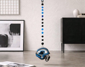 Op de afbeelding: Een zilveren voetbalhelm-ornament met blauwe en zwarte accenten, met het logo van de Carolina Panthers. Het ornament hangt aan een kralenketting van zwarte en blauwe kralen. De helm is gedecoreerd met blauwe edelstenen.