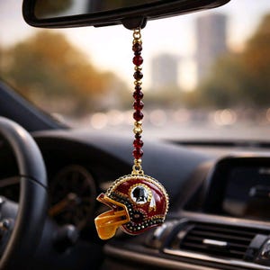 May include: A burgundy and gold football helmet car charm suspended from a rearview mirror. The helmet features a Native American profile and is decorated with red and gold beads. The charm hangs inside a car.